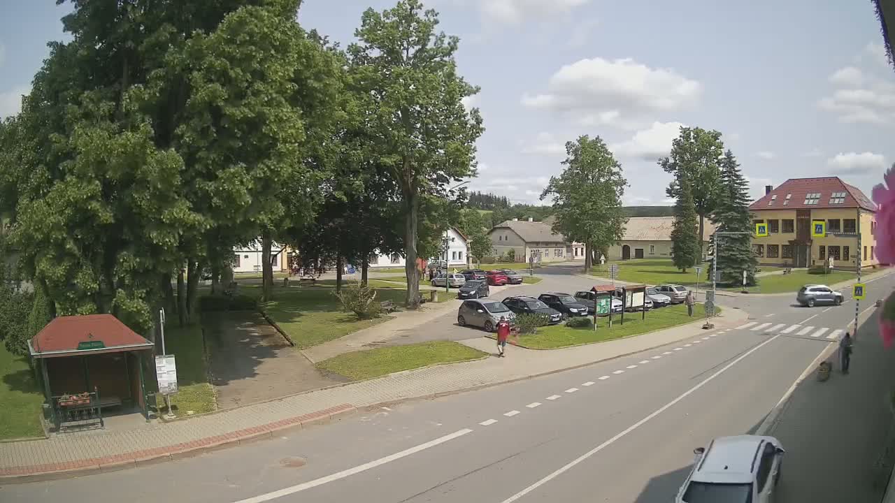 A sunny day in a town square with several parked cars, a small park with green grass and mature trees, and several buildings visible in the background.