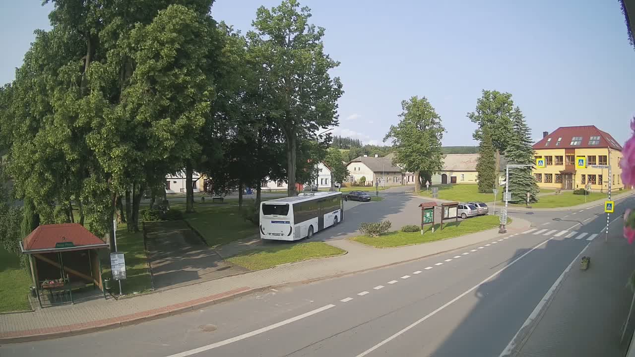 A white bus is parked in a village square on a sunny day, surrounded by buildings, trees, and a few cars.