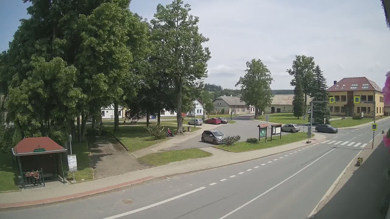 A sunny day in a village square shows several cars parked and driving near buildings, green lawns, and leafy trees.