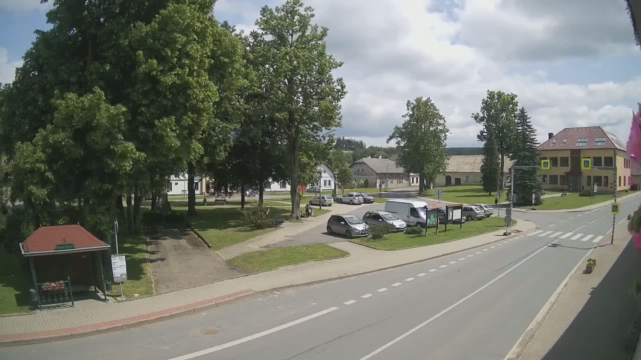 A partly sunny day shows a village square with several cars parked near a bus stop and buildings surrounding a grassy area.