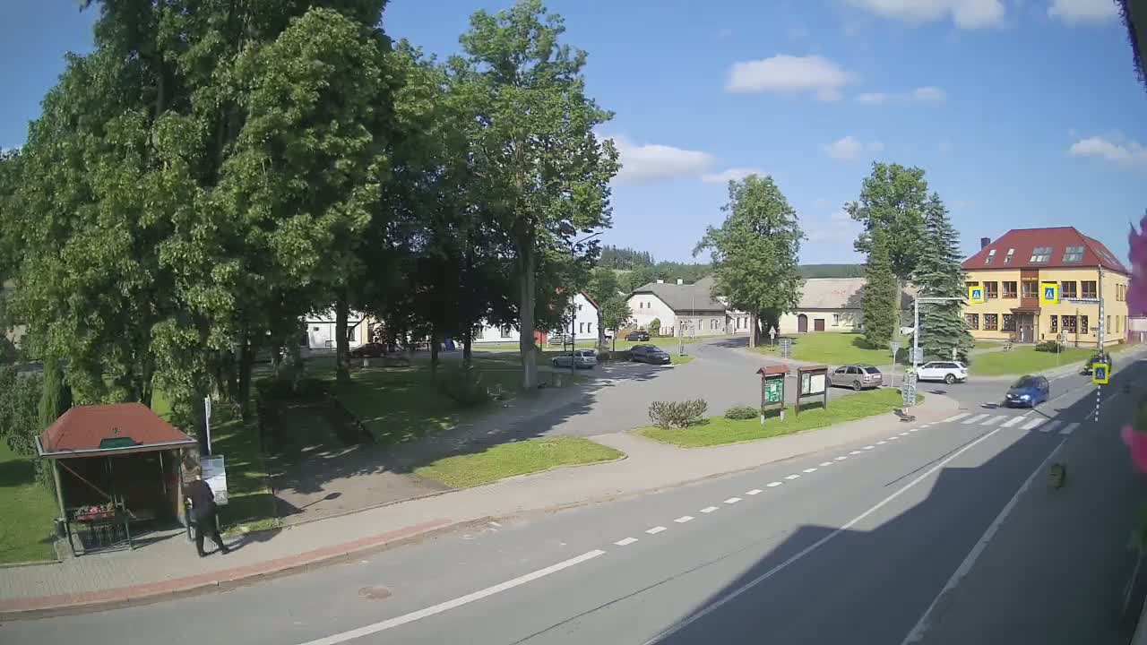 A sunny day in a village square, showing several buildings, a few cars, some trees, and a person standing near a bus stop.