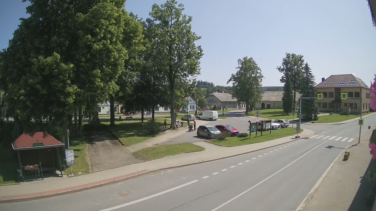 A sunny village square with several cars parked, a few people gathered near some trees, and a two-story building in the background.