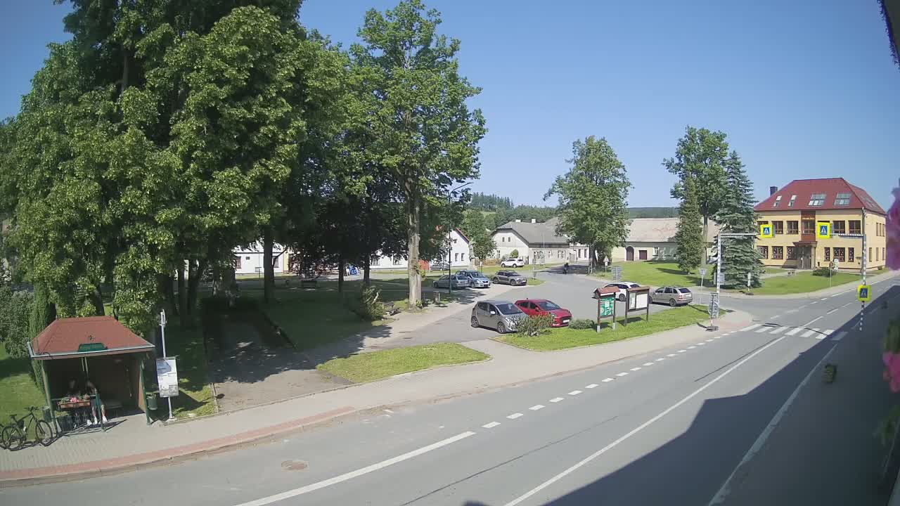A sunny day in a small town shows a village square with several parked cars, a bus stop with people waiting, and several buildings, including a two-story building with a red roof.