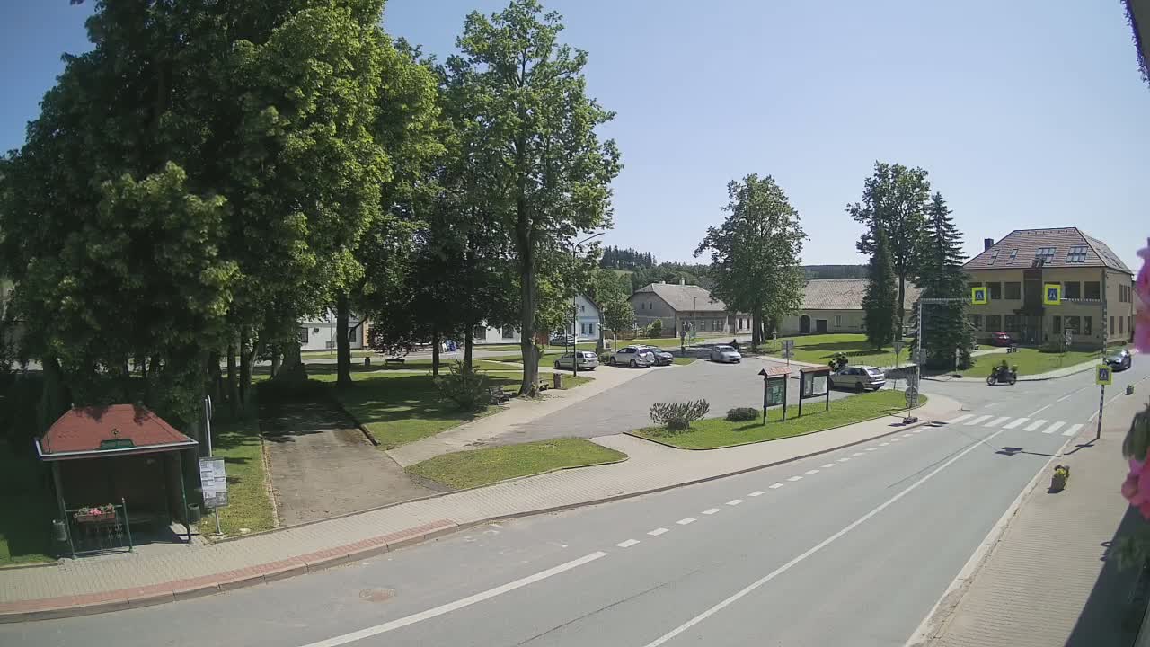 A sunny village square with a few cars parked, a small park, and buildings surrounding it, all under a clear blue sky.