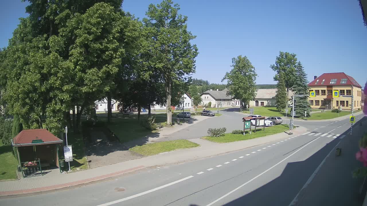 A sunny day reveals a village square with a few cars parked, several buildings, and lush green trees surrounding a bus stop.