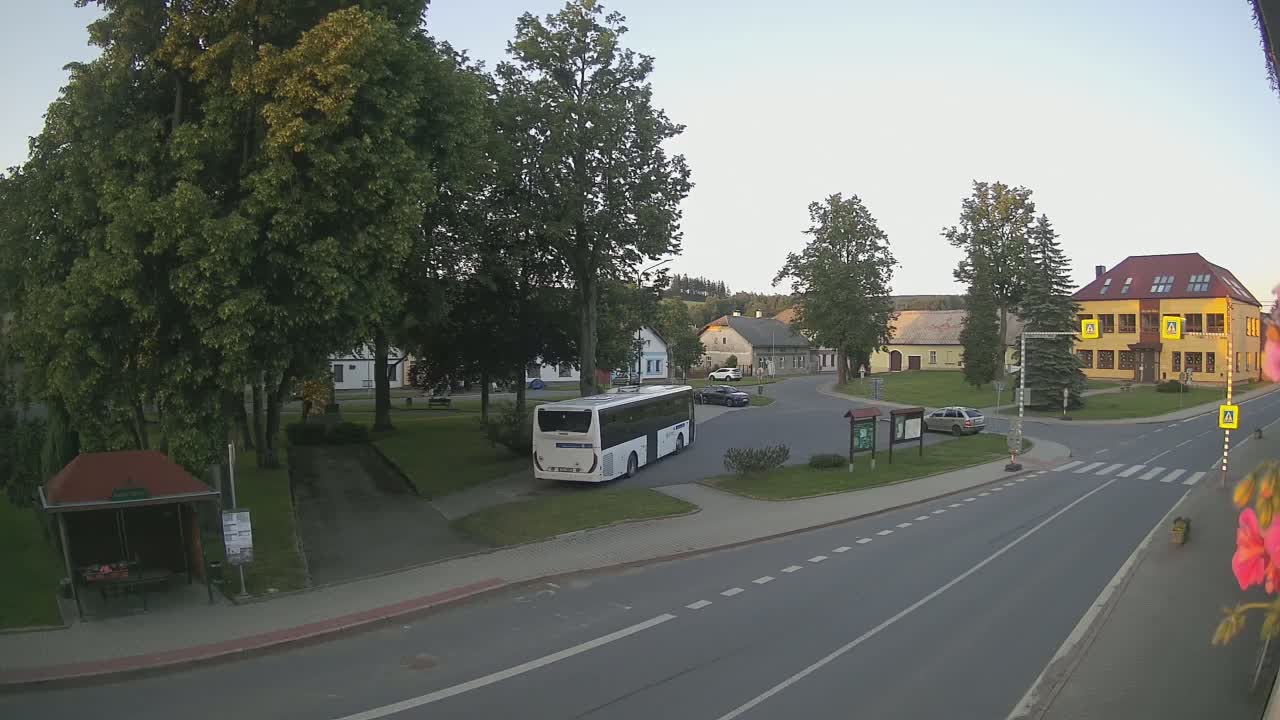 A white bus is parked in a village square on a sunny day, with several cars and buildings visible, surrounded by green trees and grass.