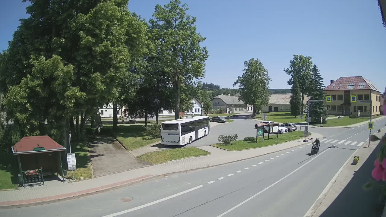 A white bus is parked in a village square on a sunny day, with a motorcyclist riding on a road nearby.