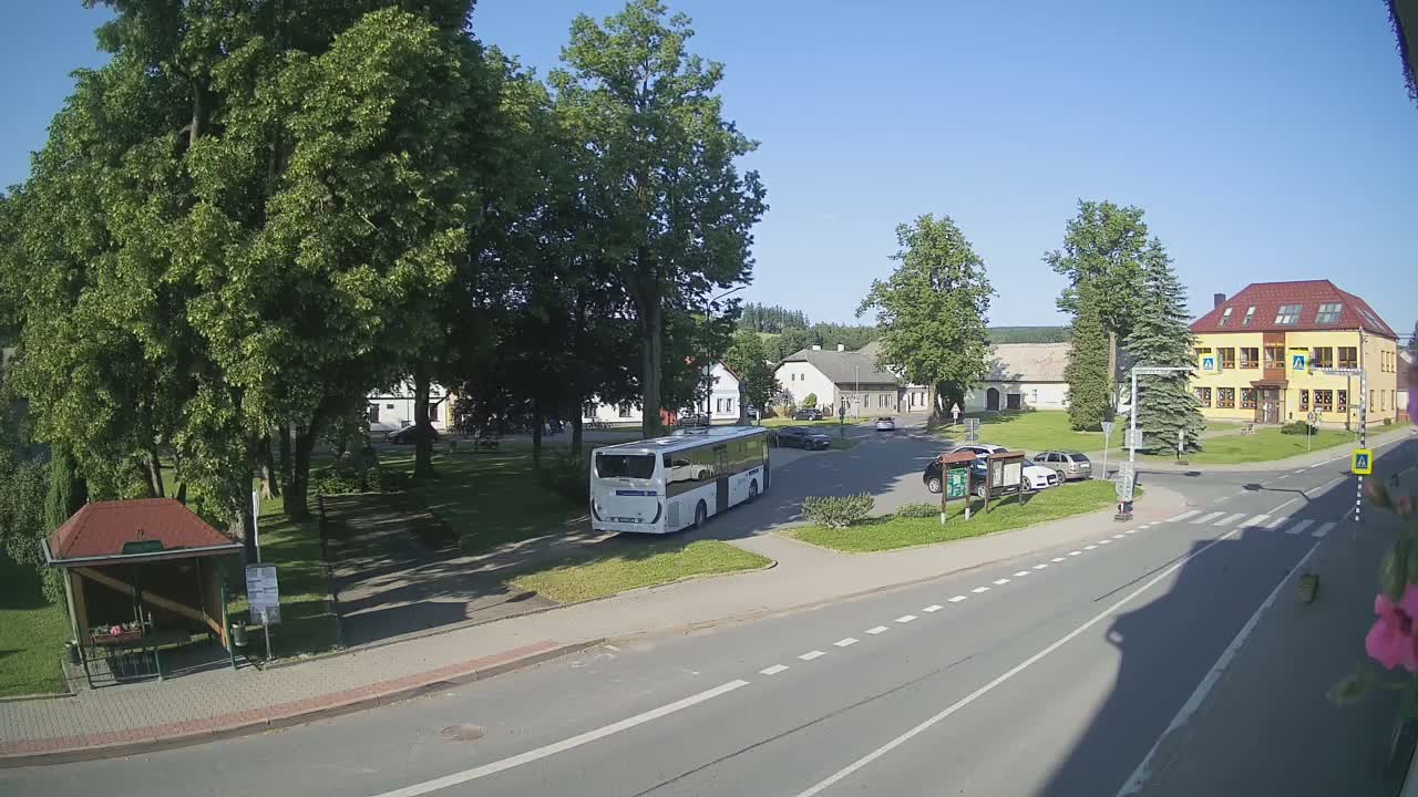 A white bus is parked in a village square on a sunny day, near a bus stop and several buildings, with cars parked nearby.