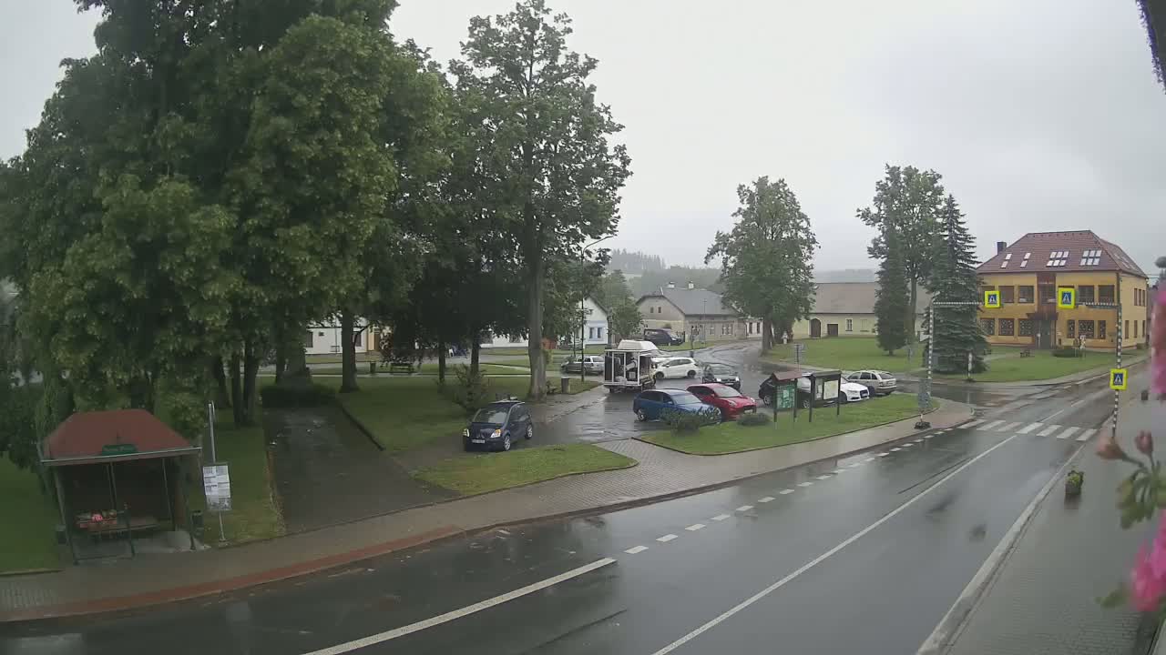A rain-soaked town square with several cars parked near a bus stop and several buildings, surrounded by lush green trees.