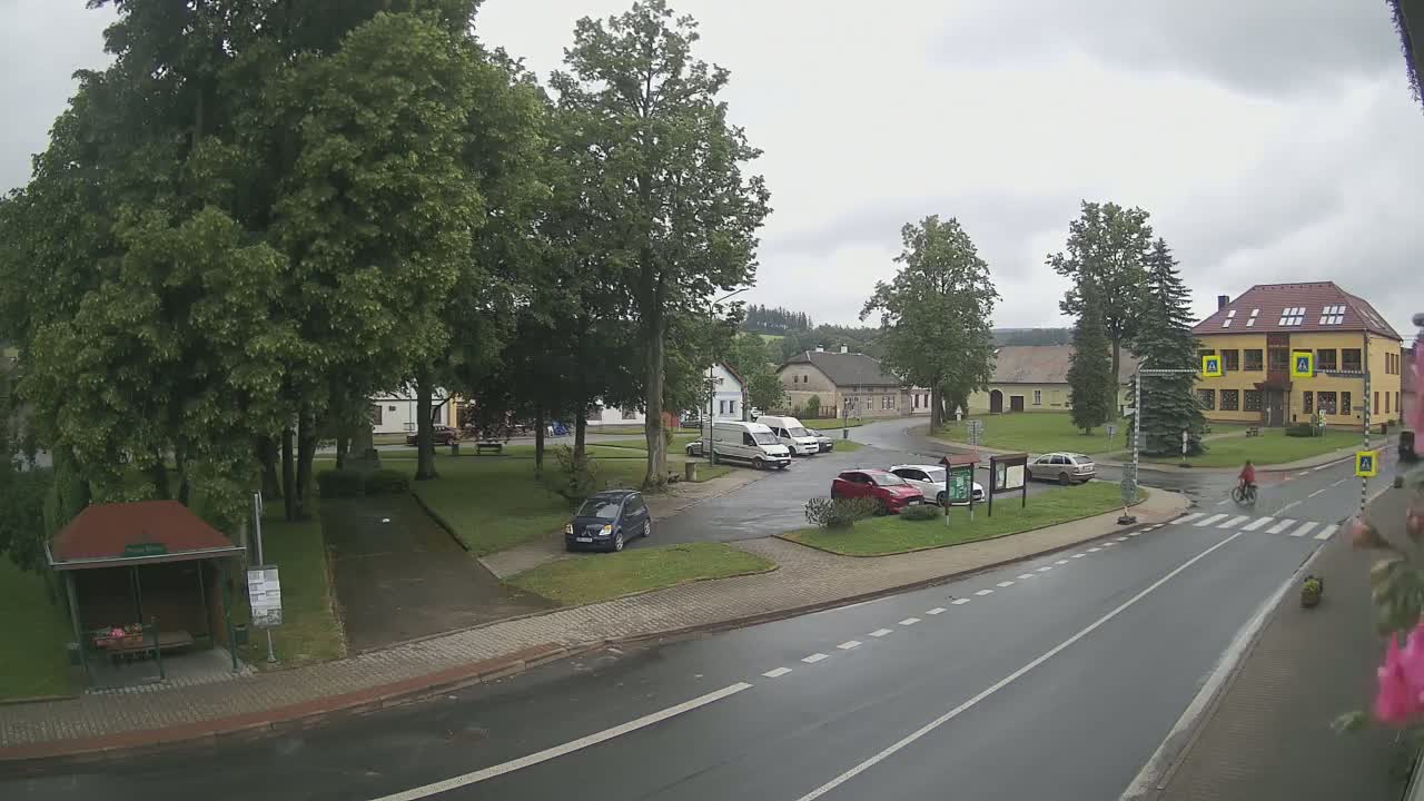 A small town square with several cars parked, a bus stop, and a two-story building, all under a cloudy sky.