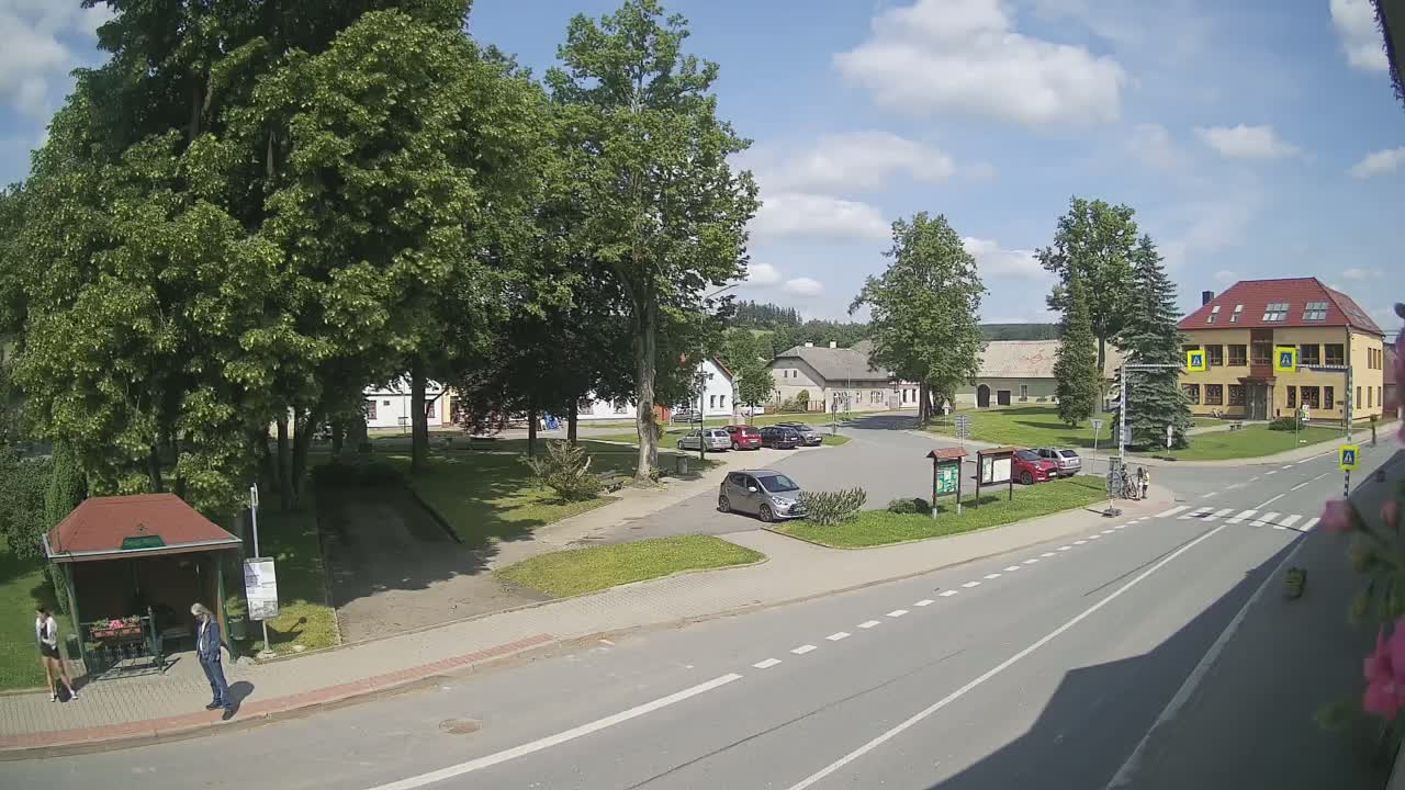 A sunny day in a small town square shows a few cars parked, people walking near a bus stop, and several buildings surrounding a grassy area.