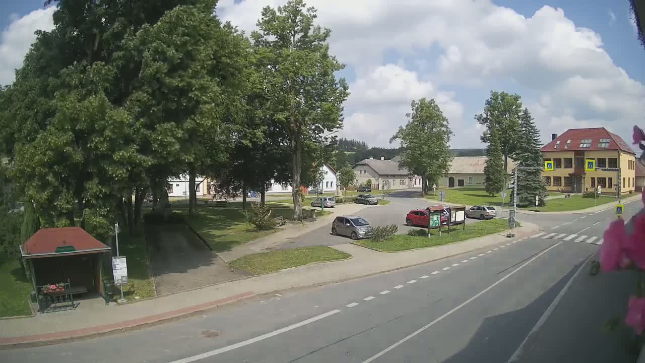 A partly sunny day reveals a village square with several parked cars, buildings, and lush green trees surrounding it, all near a paved road.