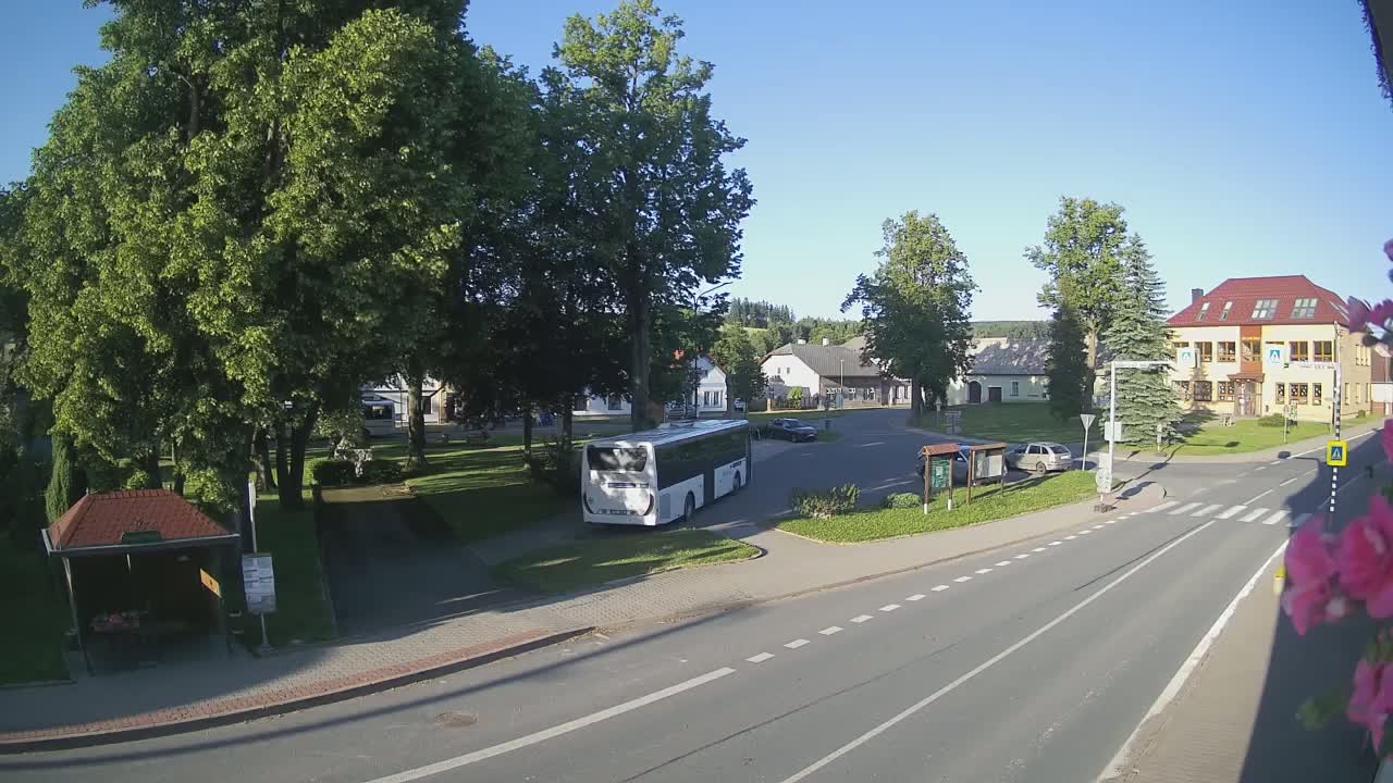 A white bus is parked in a village square on a sunny day, near a small park and several buildings.
