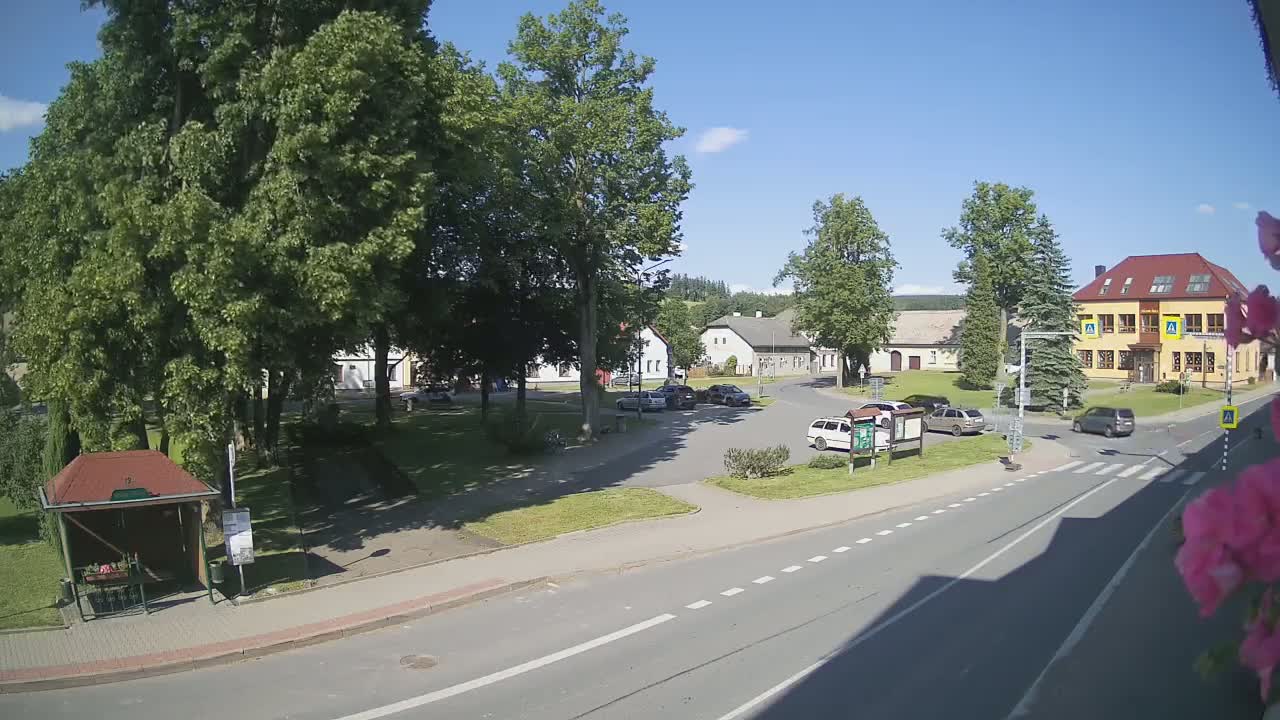 A sunny day reveals a village square with several cars parked, a bus stop, and a few buildings surrounded by lush green trees.