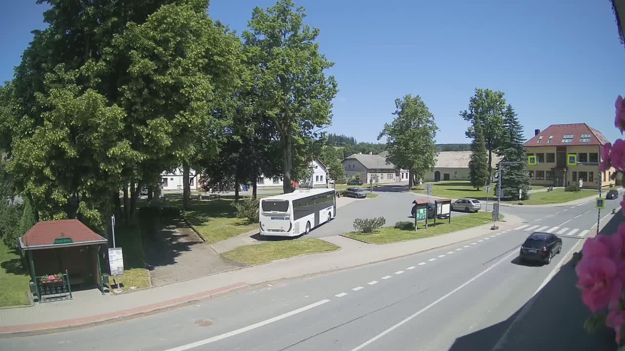 A white bus is parked in a village square on a sunny day, with several cars on the surrounding roads and several buildings nearby.
