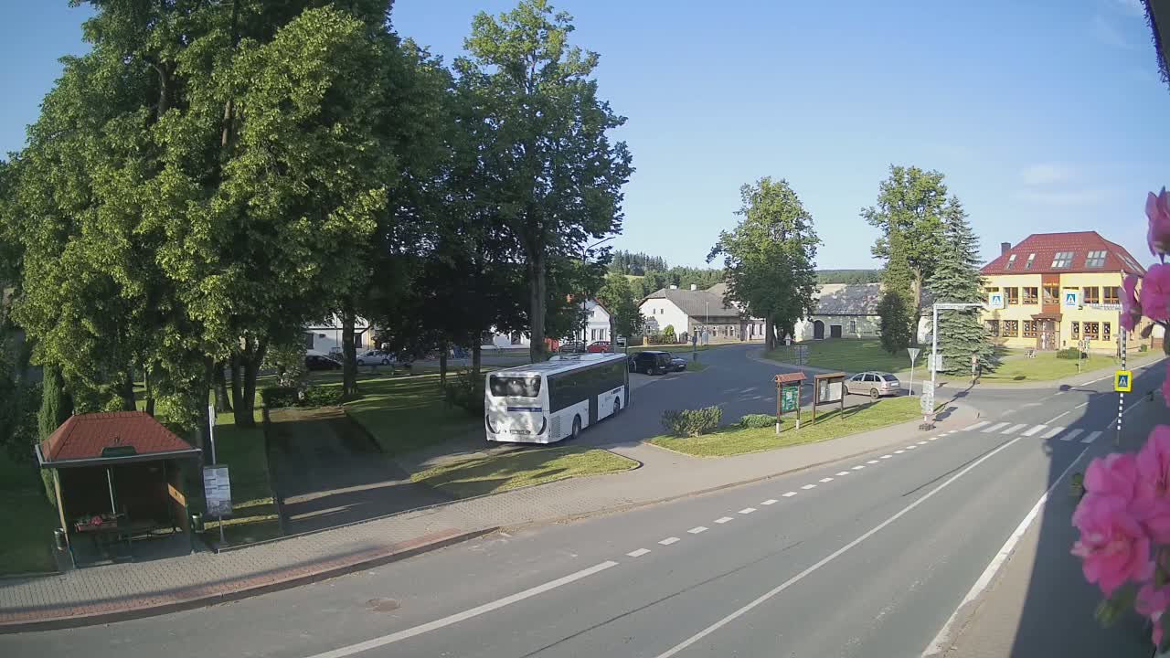A white bus is parked in a village square on a sunny day, surrounded by buildings, trees, and a few cars.