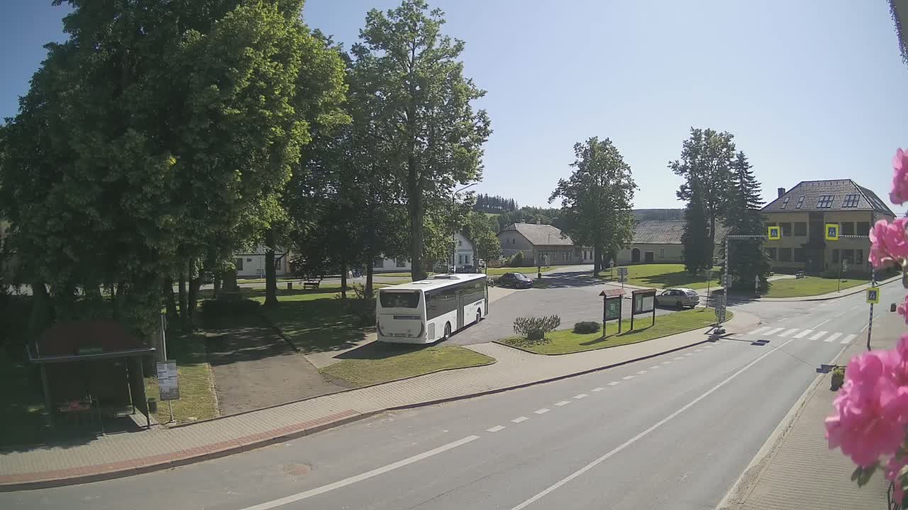 A white bus is parked in a village square on a sunny day, with several buildings and green spaces surrounding it.