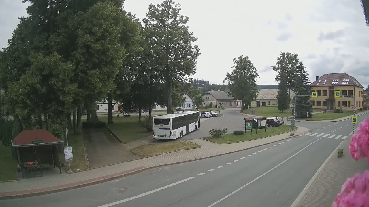 A white bus is parked in a village square on an overcast day, surrounded by buildings, trees, and a few cars.