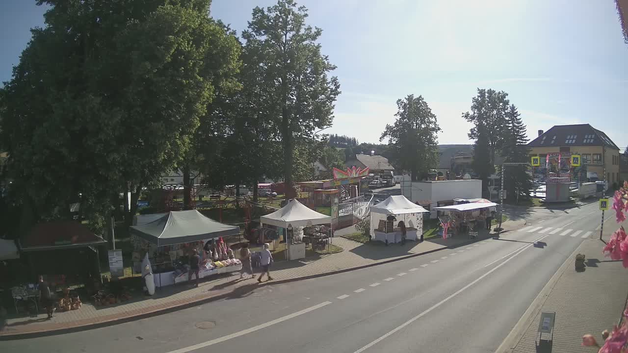 A sunny outdoor scene shows a street with market stalls and a small fairground ride, situated near several buildings and large trees.