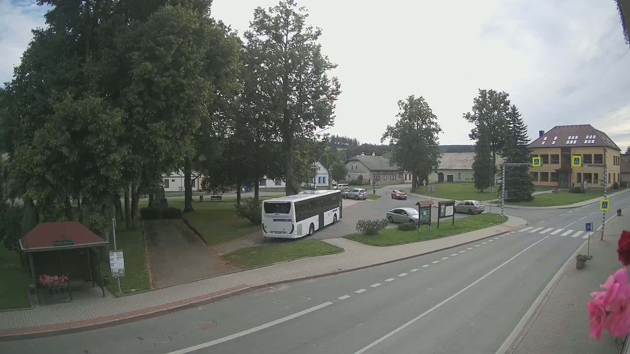 A white bus is parked in a small village square under a partly cloudy sky, with several cars parked nearby and buildings lining the square.