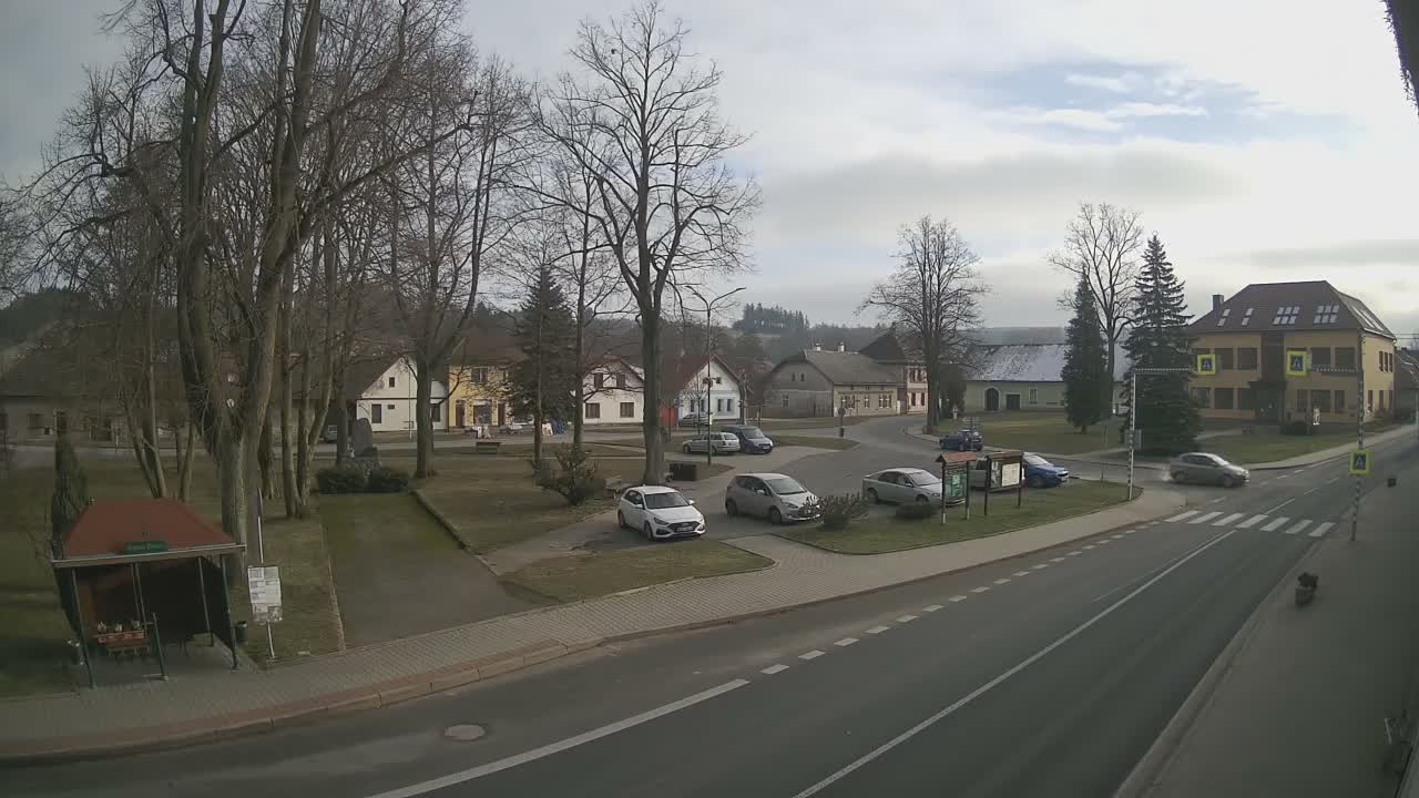 An overcast day reveals a quiet village square with bare trees, a bus stop, and parked cars alongside a road intersection, surrounded by various buildings and distant hills.
