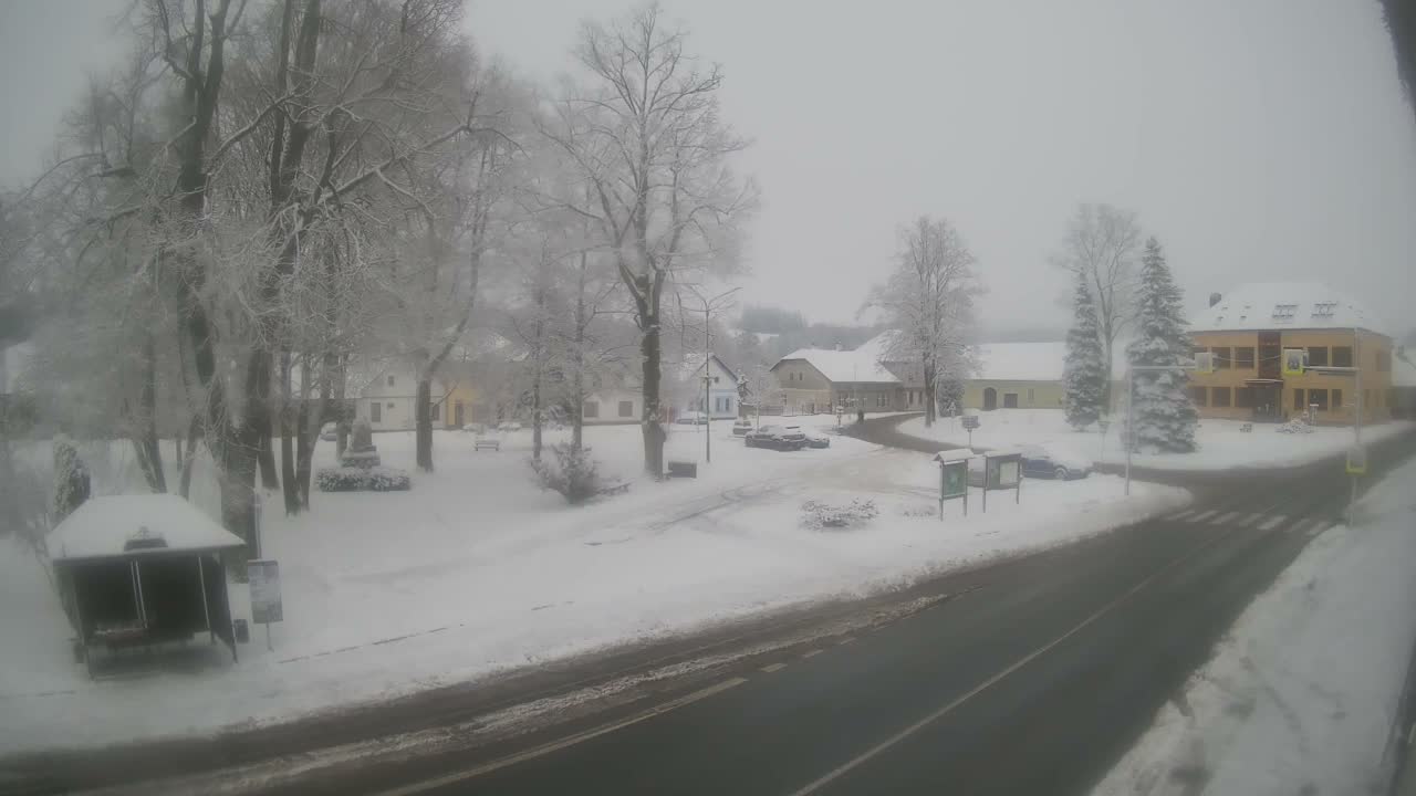 A snow-covered village square and surrounding area are depicted under a hazy, overcast sky, with trees, rooftops, and ground blanketed in white, and a partially cleared road cutting through the scene.