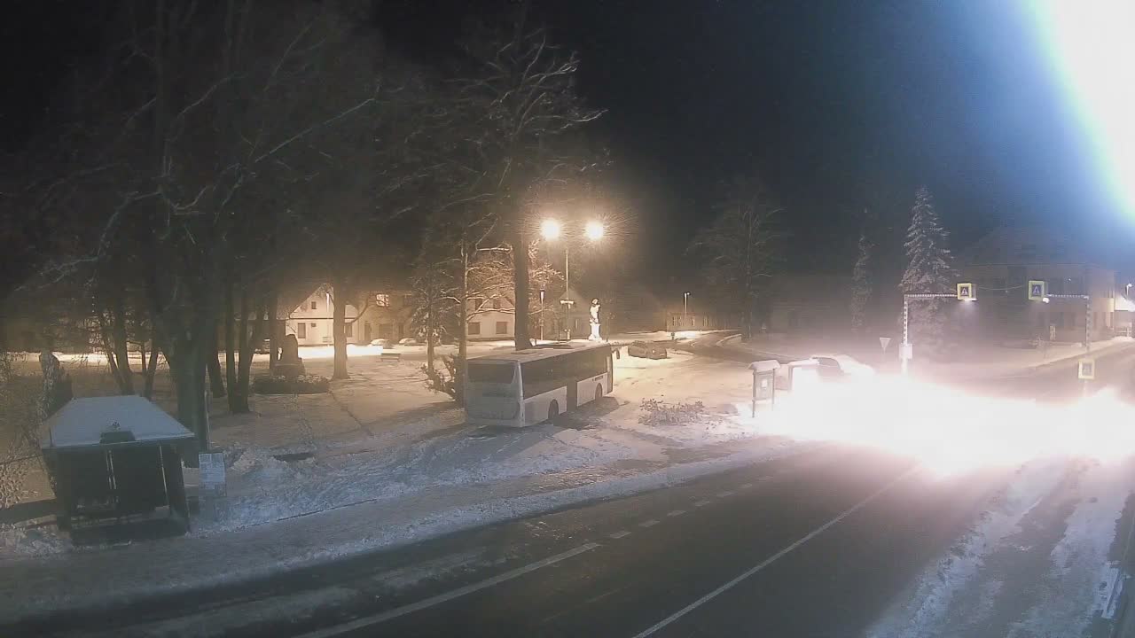 The image shows a snowy village street at night, with a bus parked among snow-covered trees and buildings, while bright vehicle headlights pierce through falling snow.