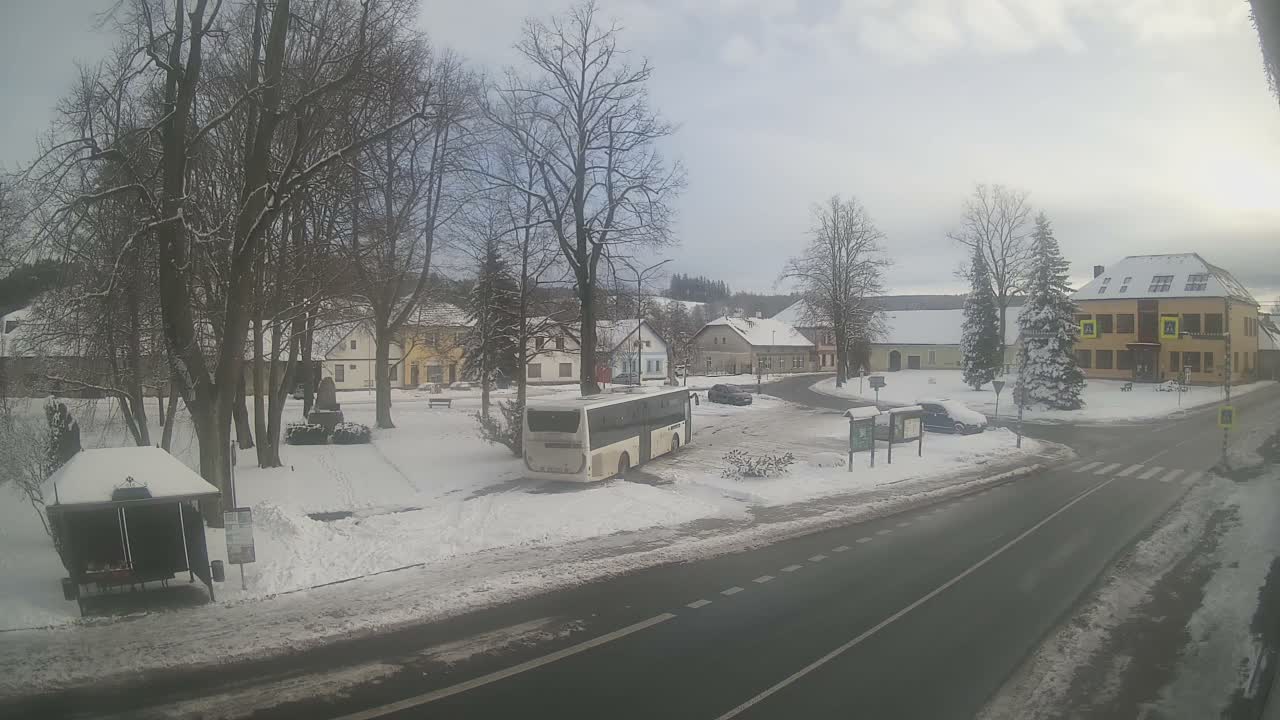 The image displays a snow-covered village street on an overcast winter day, featuring bare trees, houses with snowy roofs, a parked white bus, and a partially cleared road.