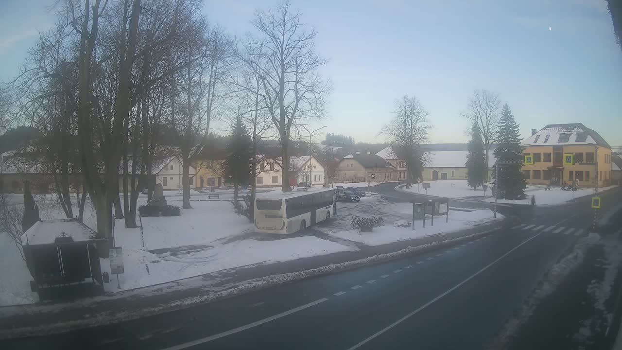 The image displays a snow-covered village street on an overcast winter day, featuring bare trees, houses with snowy roofs, a parked white bus, and a partially cleared road.