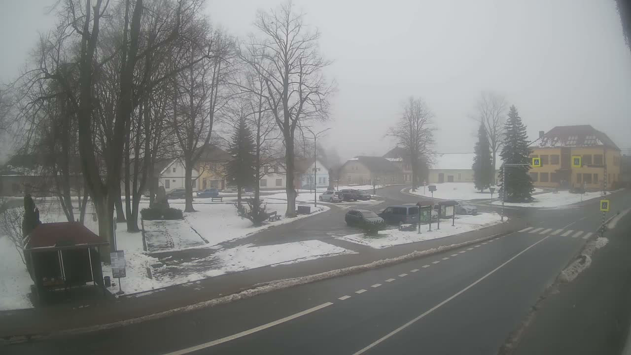 A hazy winter scene captures a snow-dusted village square on a foggy day, featuring bare trees, parked cars, and several buildings surrounding a road intersection.