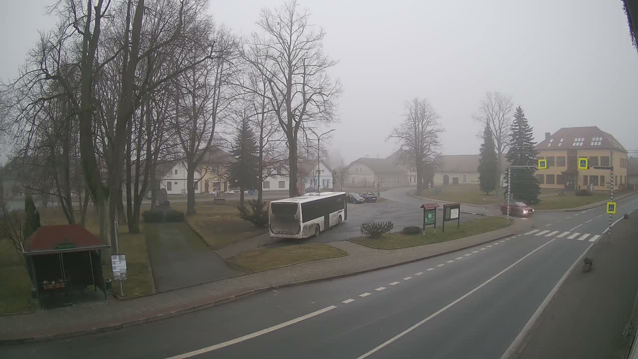 A hazy winter scene captures a snow-dusted village square on a foggy day, featuring bare trees, parked cars, and several buildings surrounding a road intersection.