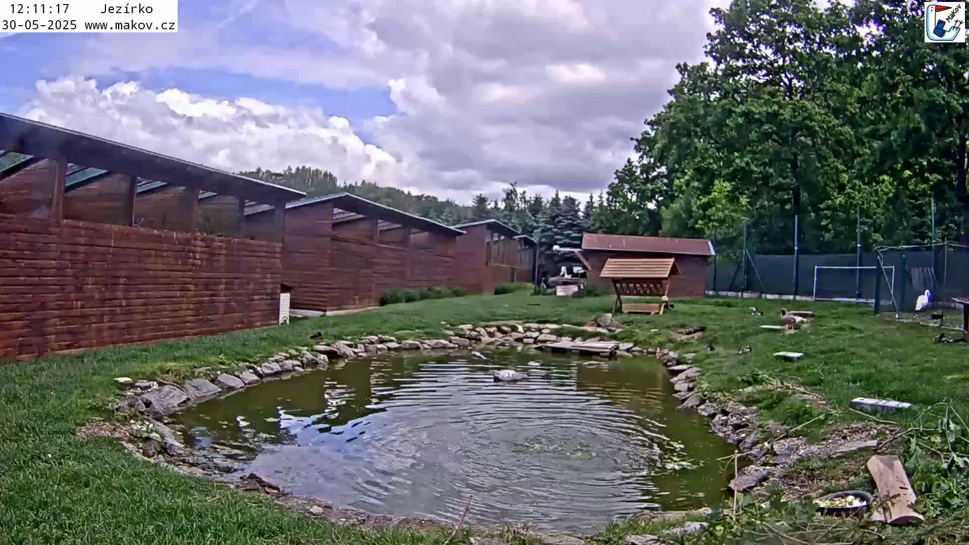 A small pond with several birds near a wooden building complex under a partly cloudy sky.