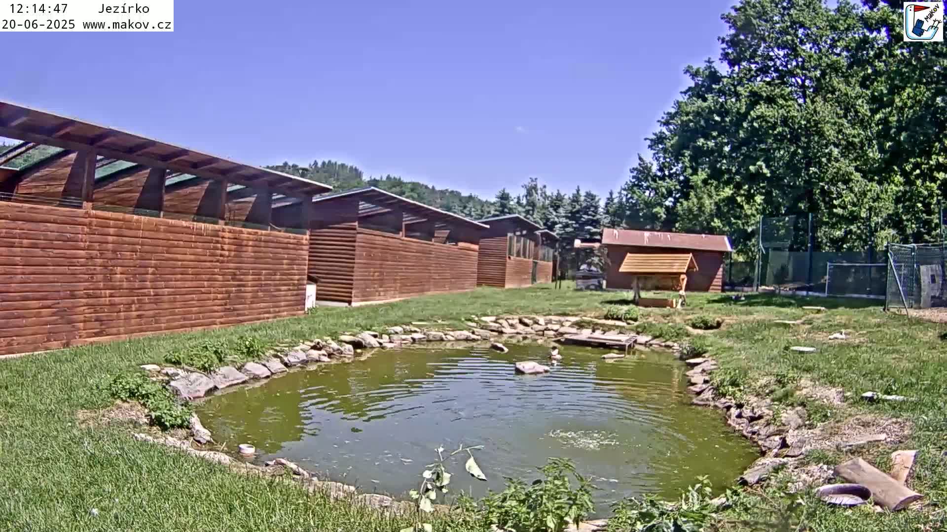 A row of brown wooden structures surrounds a small pond under a clear blue sky.