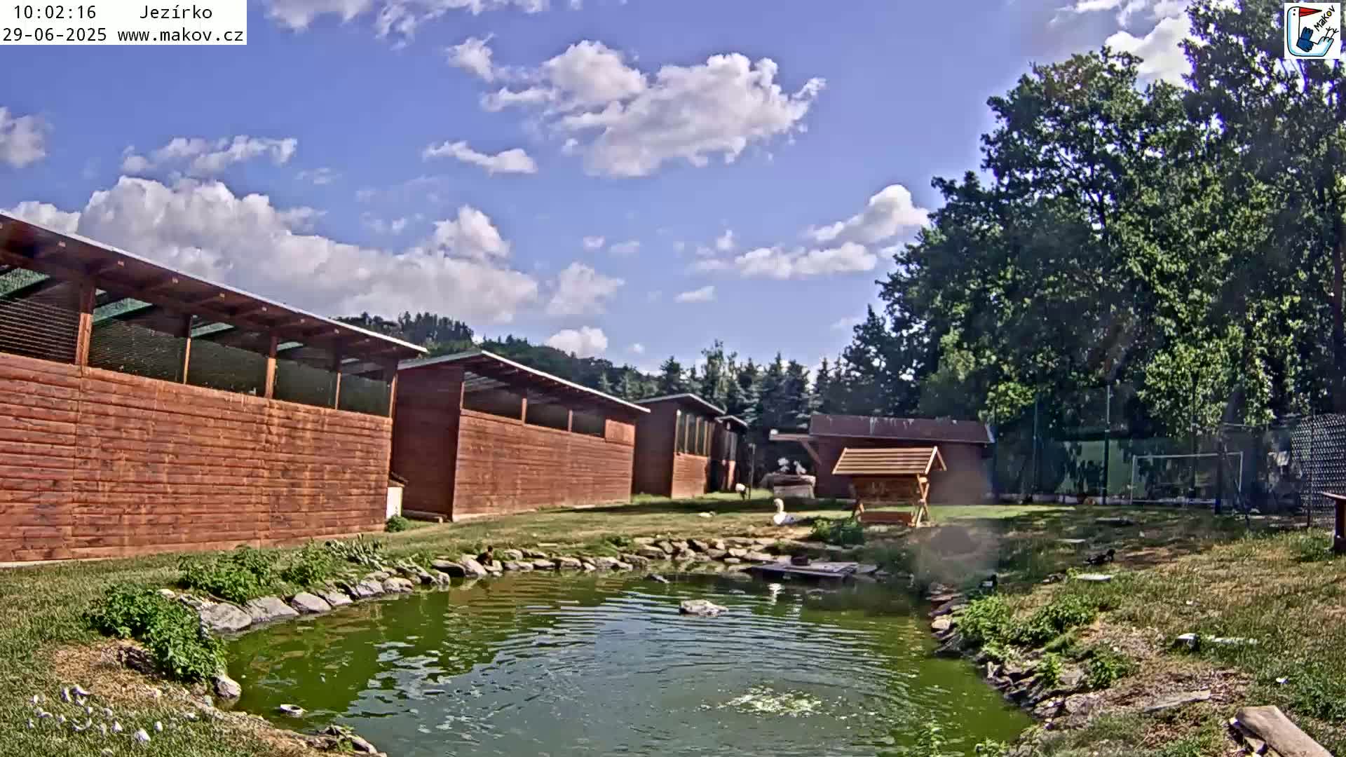 A small pond sits in front of several wooden buildings under a partly cloudy sky.