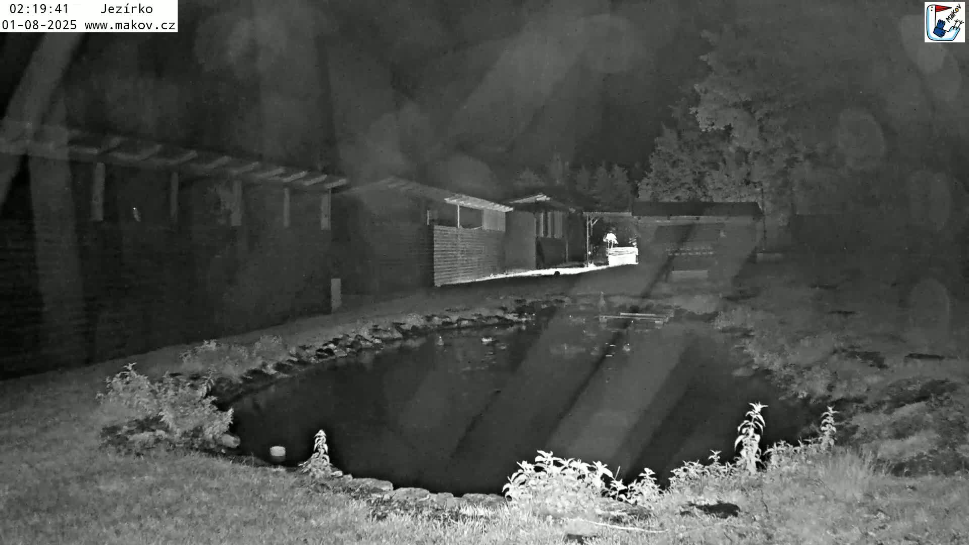 A nighttime, grayscale image shows a small pond next to a long building under a dark sky.