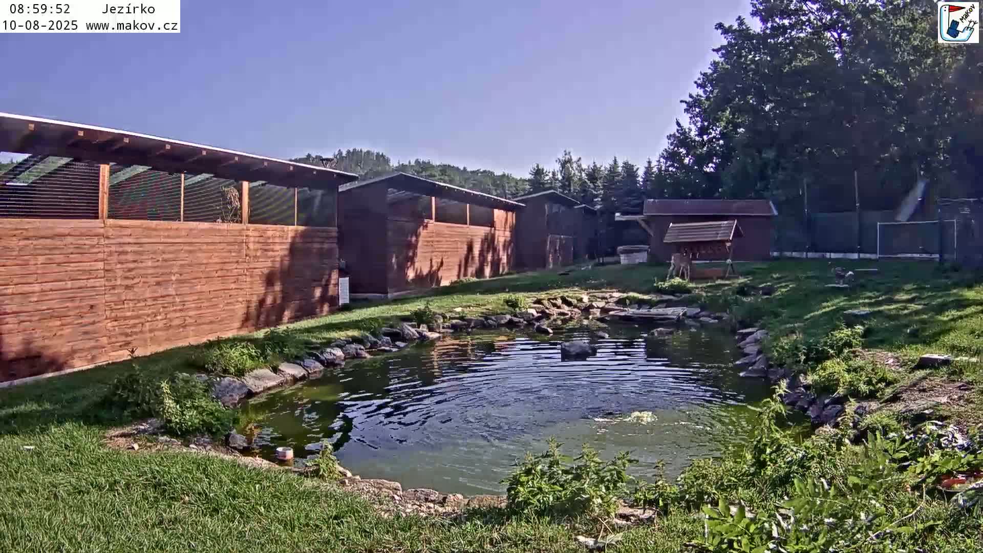 A small pond sits in front of several wooden structures under a clear, sunny sky.