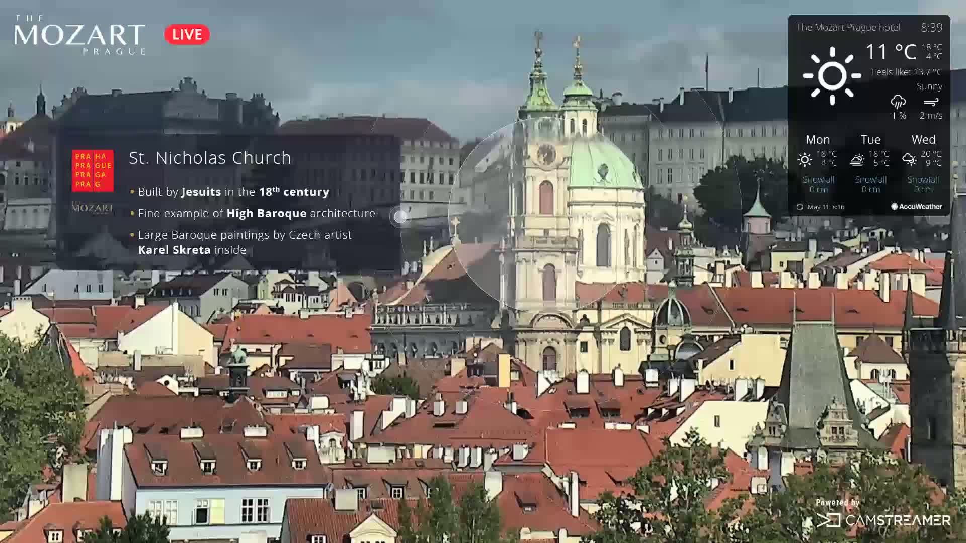 A sunny cityscape view shows a large Baroque church among many red-tiled rooftops.