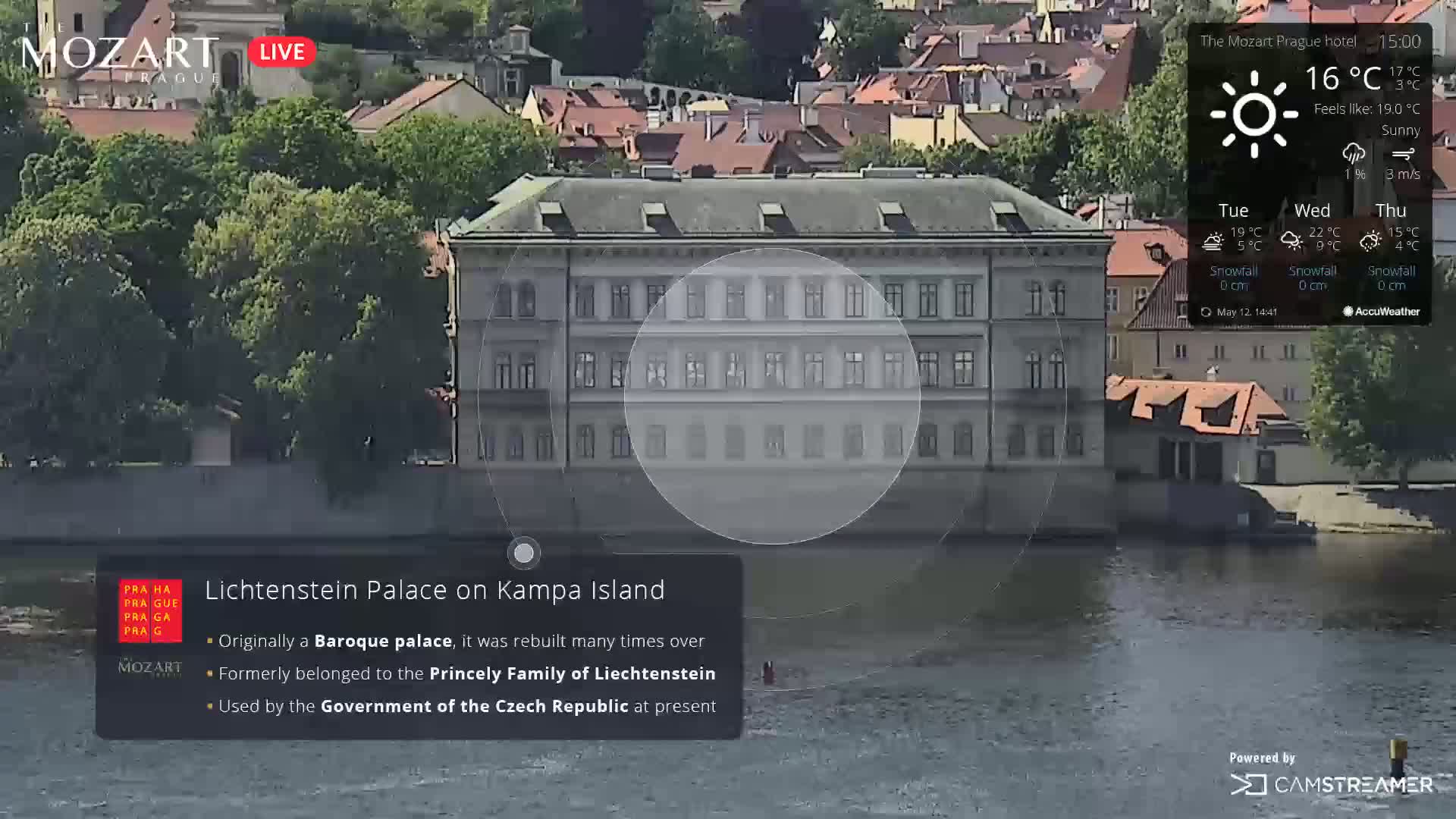 A large Baroque palace sits on the bank of a river on a sunny day.