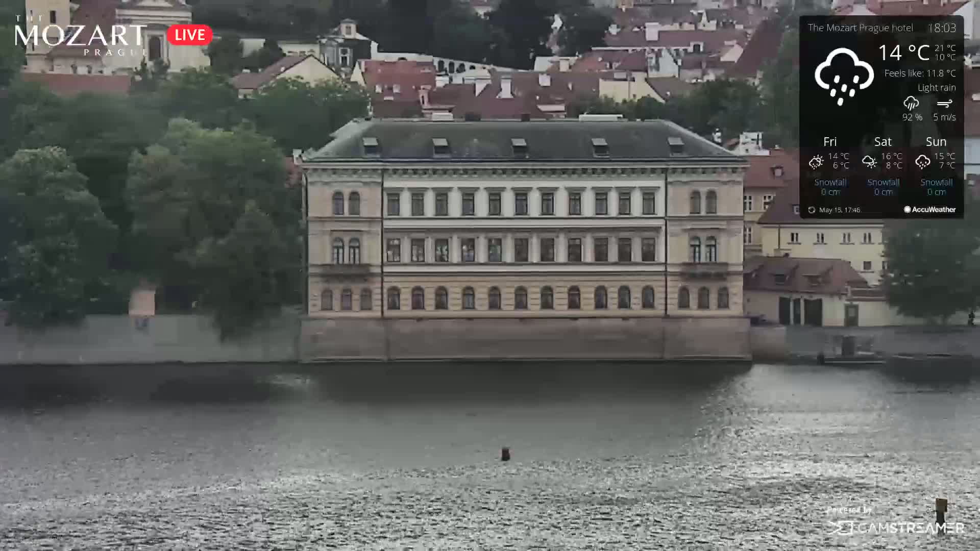 A large, multi-story building sits on the bank of a river under a light rain.
