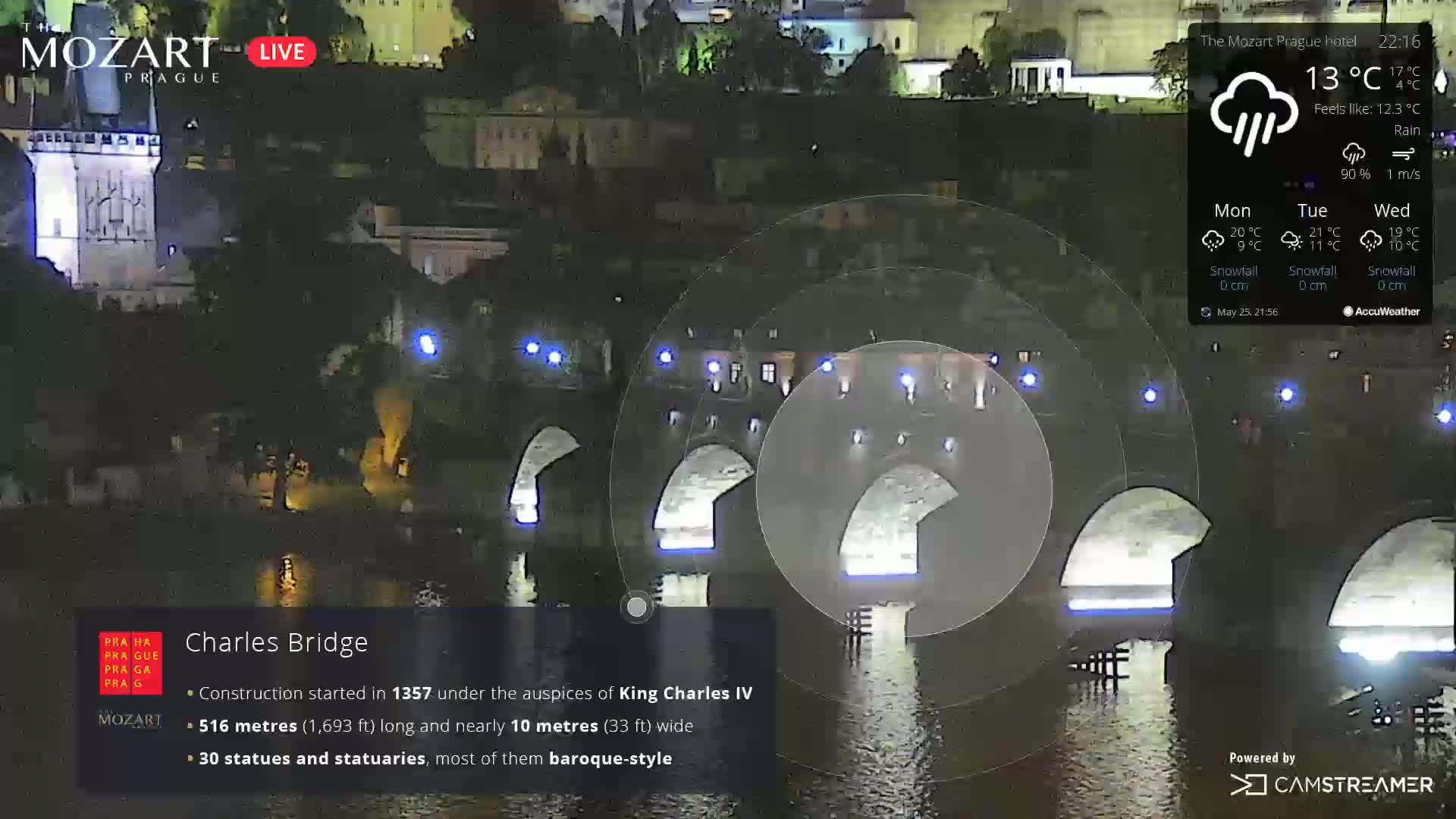 A nighttime view of a stone bridge illuminated with blue lights under rainy conditions.