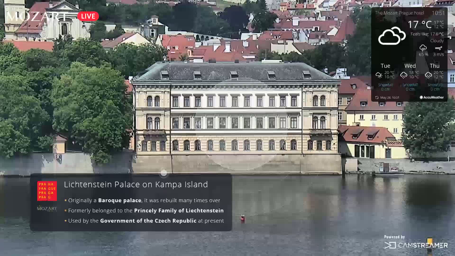 A large Baroque palace sits on a riverbank under cloudy skies, with other buildings and lush green trees visible in the background.