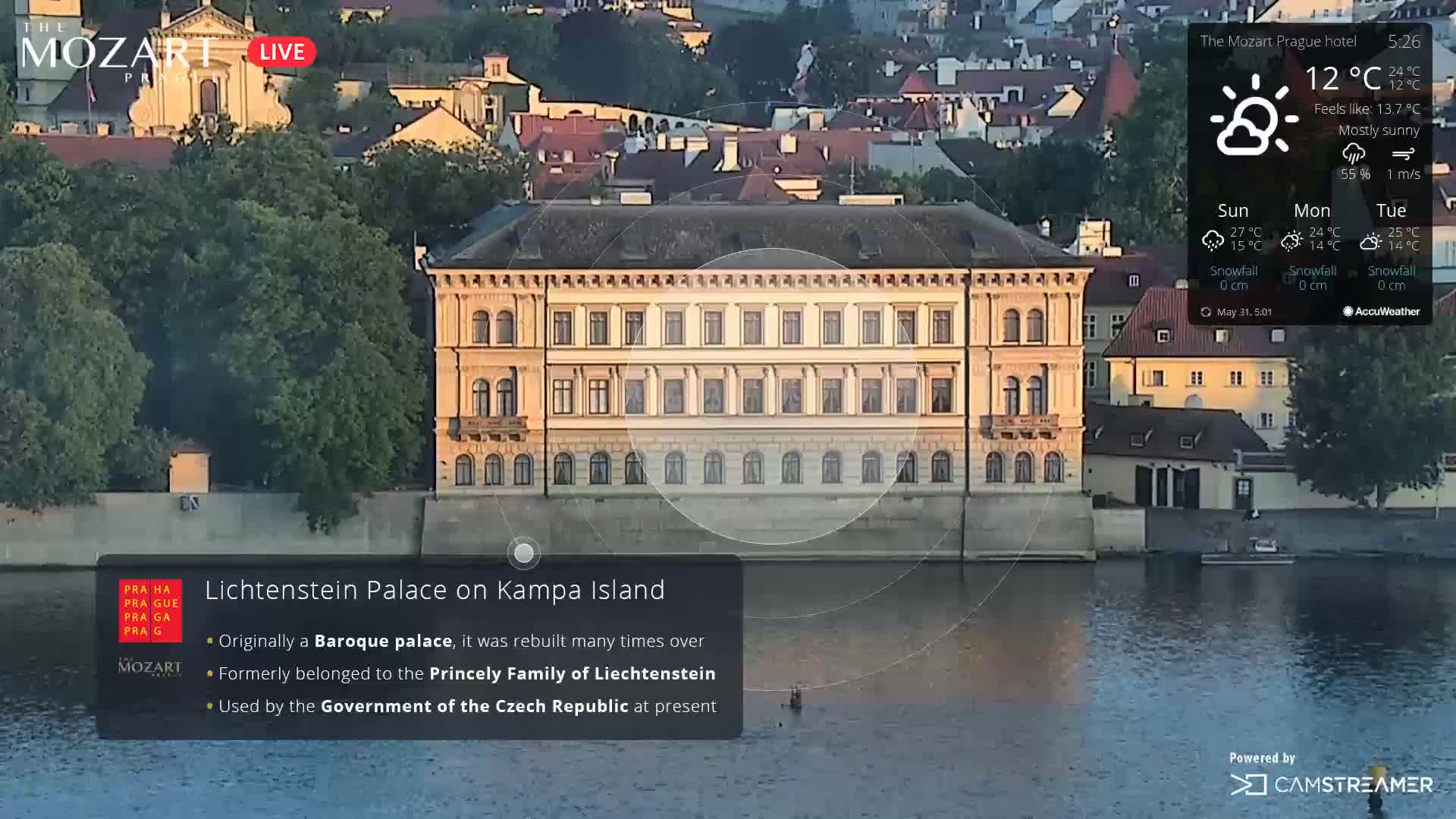 A large, pale yellow Baroque palace sits on a riverbank under a mostly sunny sky.