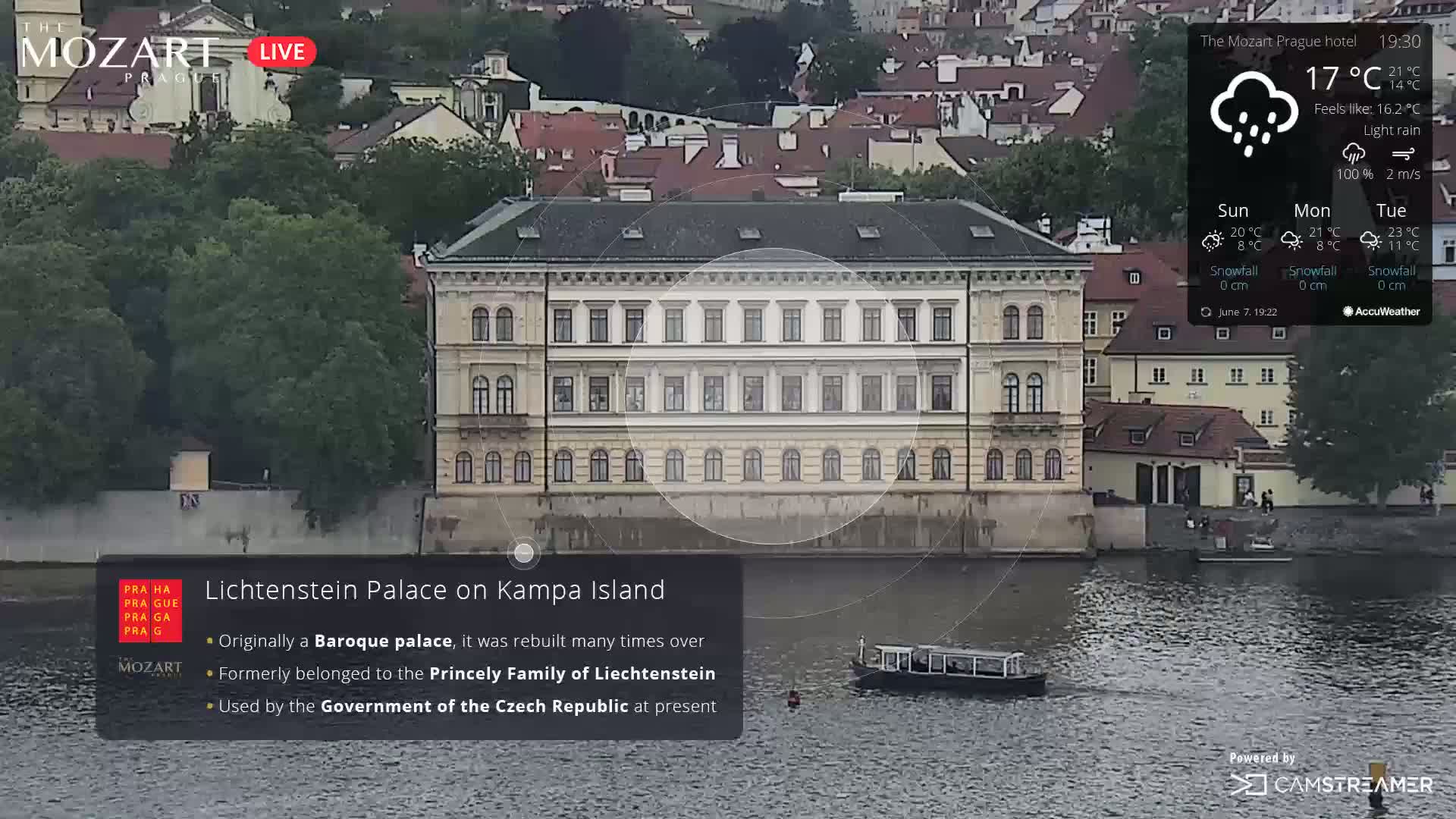 A large Baroque palace sits on a riverbank in light rain, with a small boat on the water in front of it.
