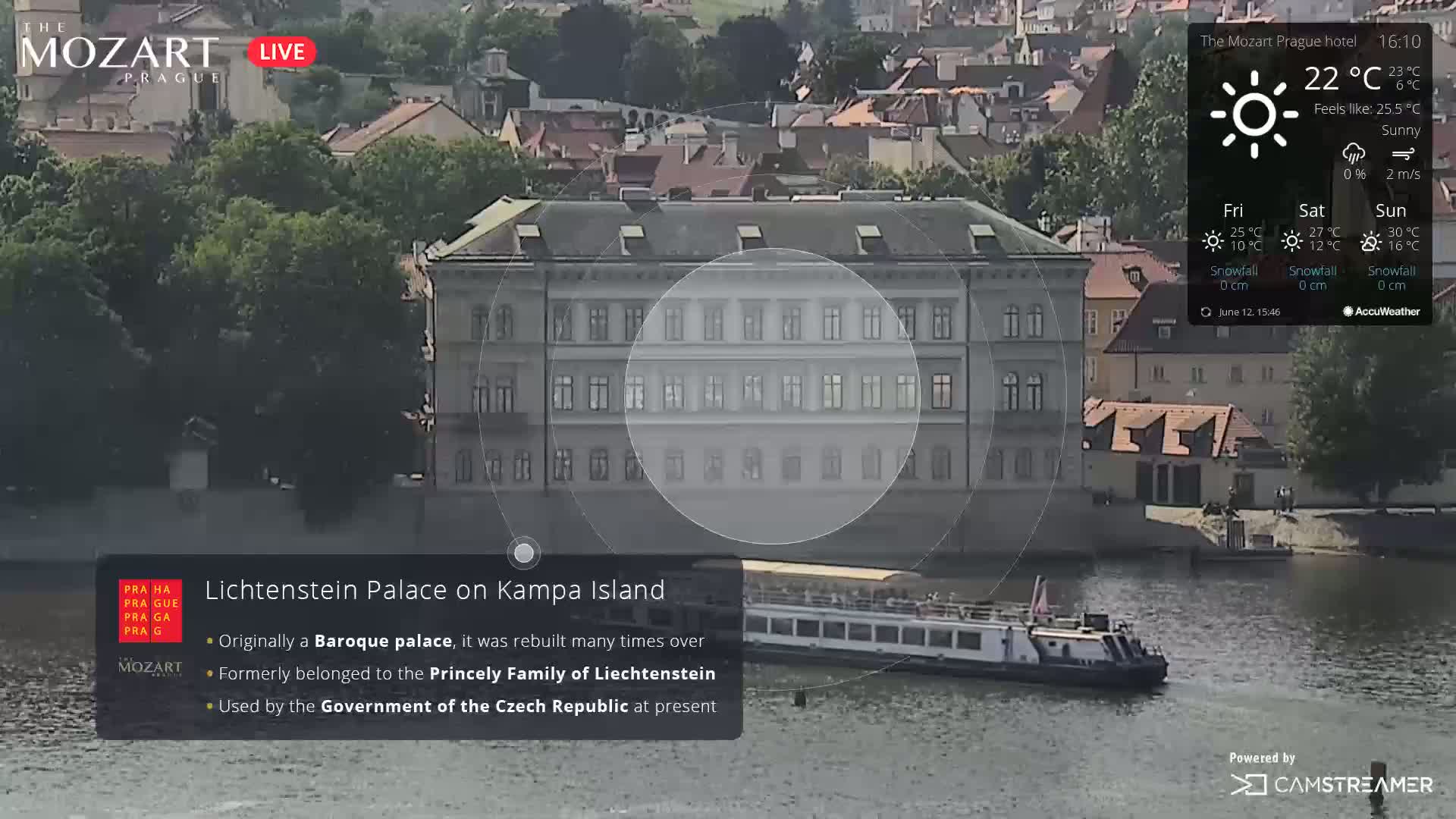 A large, light-colored palace sits on an island in a river on a sunny day, with a tour boat in the water below.