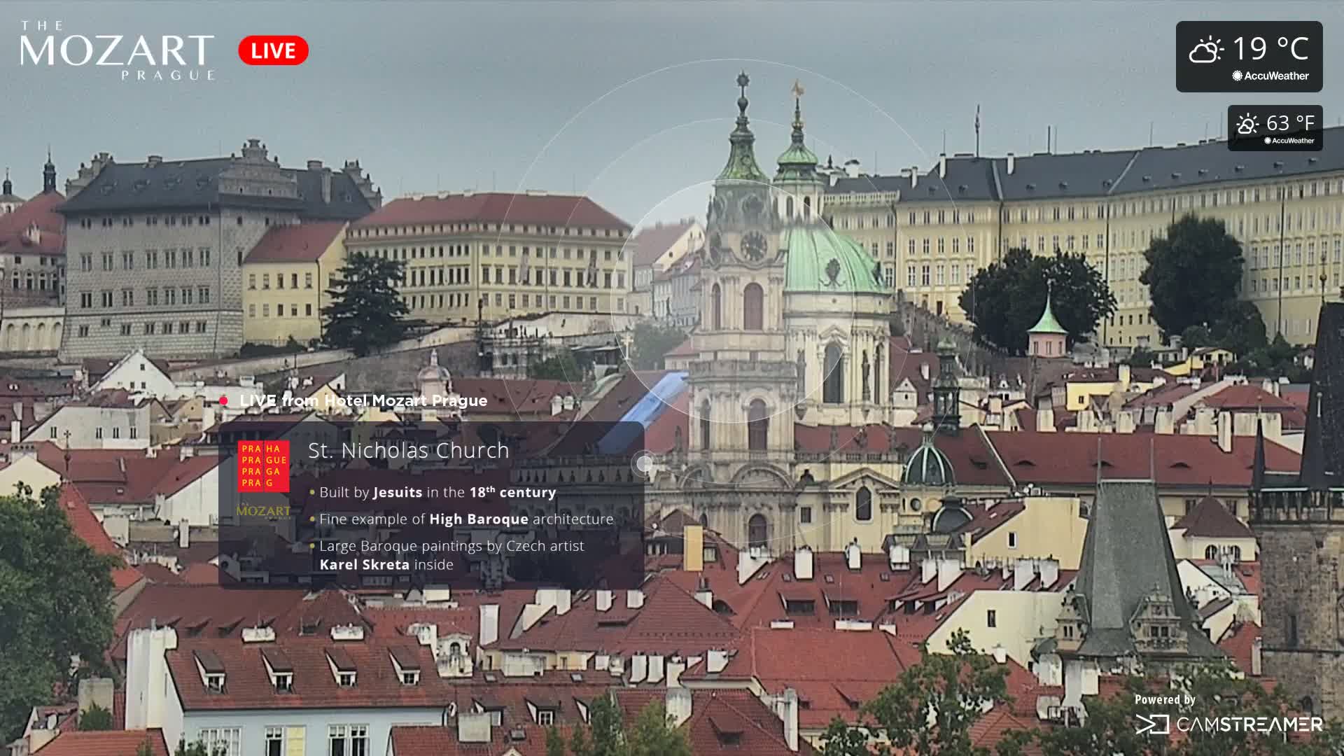 A high-angle view shows a cityscape of red-tiled roofs and buildings, prominently featuring St. Nicholas Church under a partly cloudy sky.