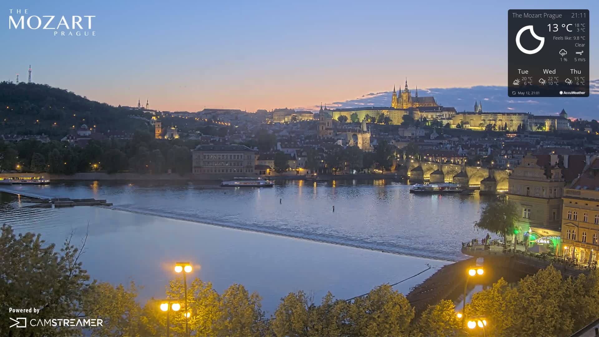A twilight view of Prague, featuring a river with a bridge and the city's illuminated buildings and castle in the background, under clear skies.
