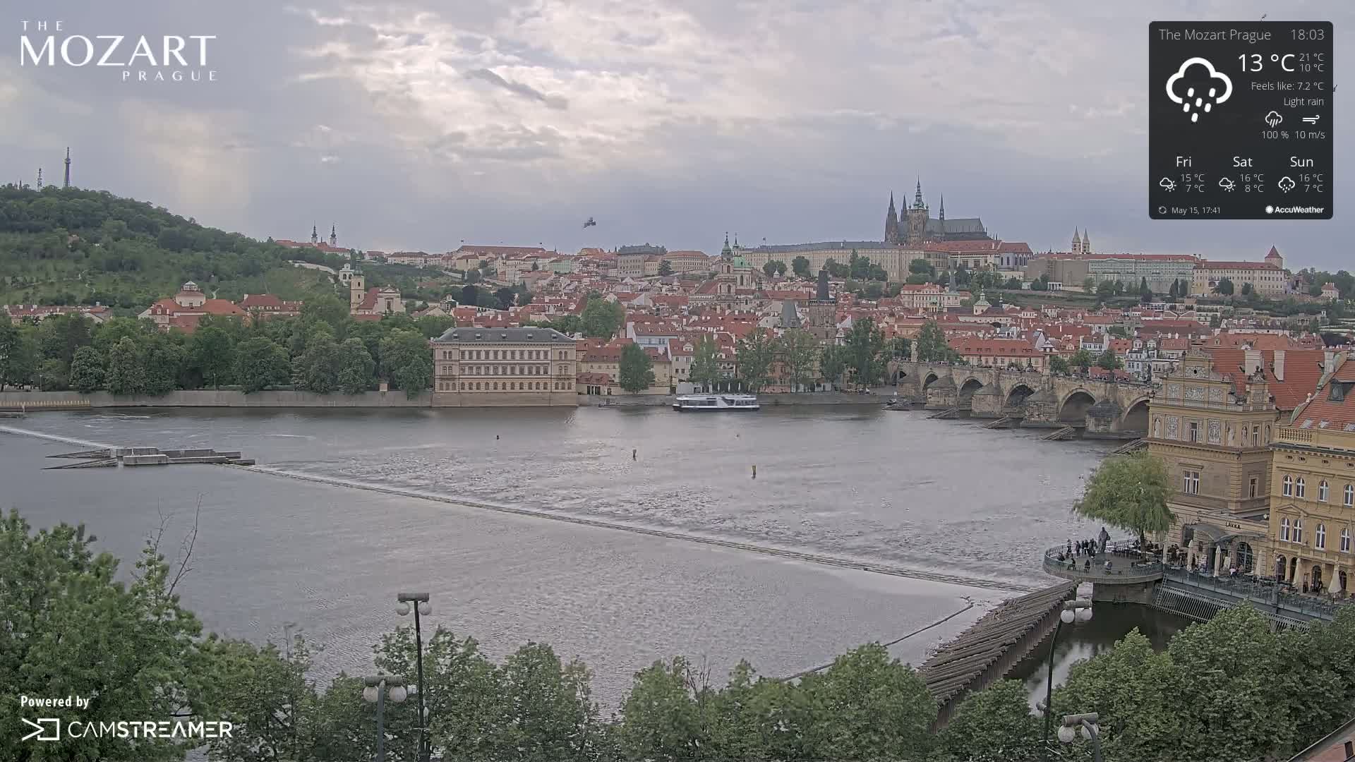 A river flows beneath a bridge in a city with buildings and trees, under a lightly overcast sky with light rain.