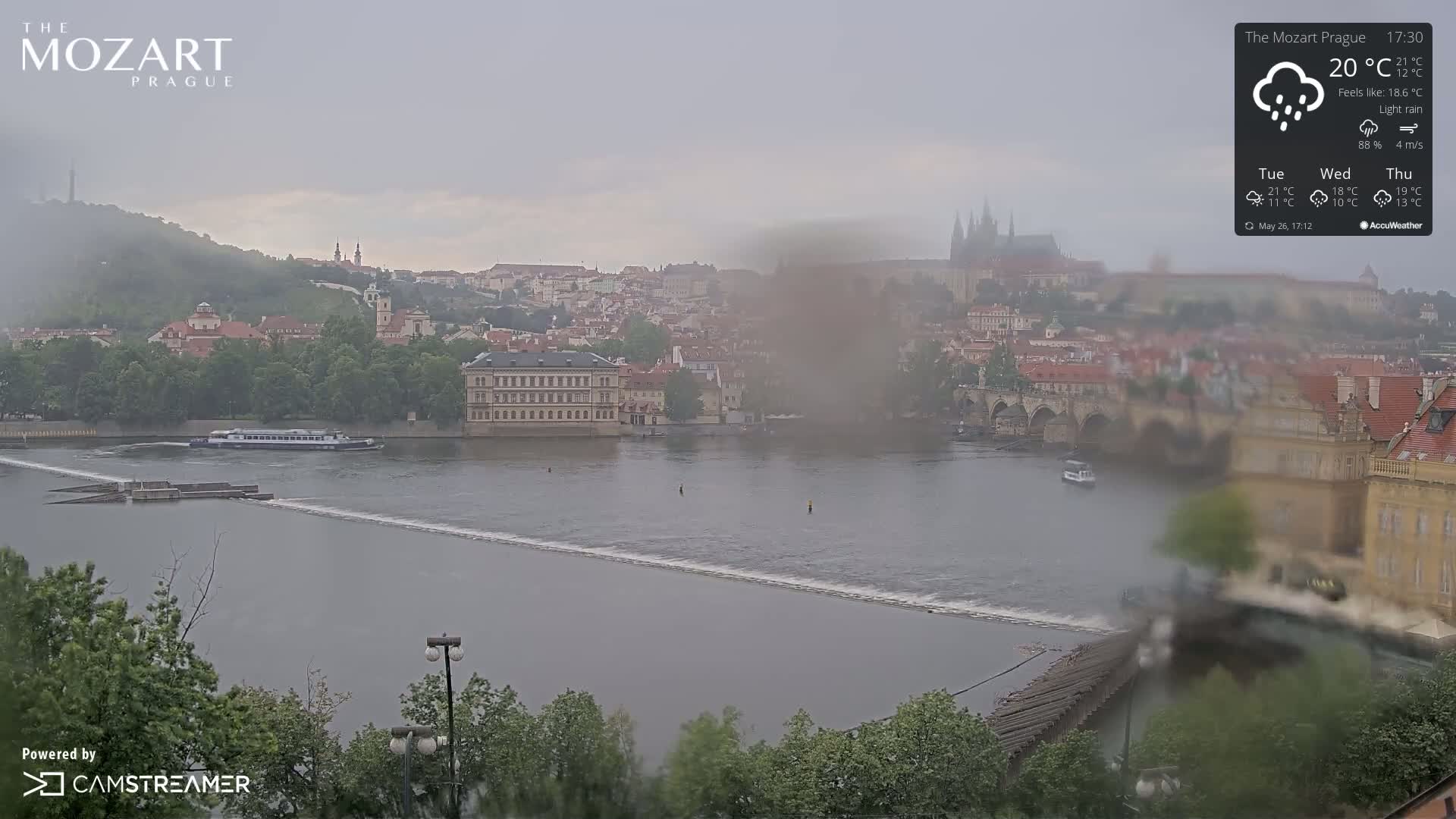 A river flows past a city with numerous buildings and a bridge, under a light rain and overcast sky.