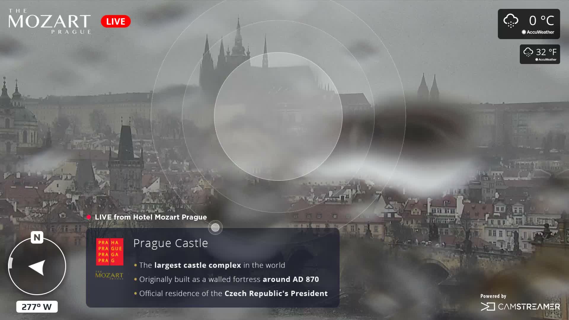 The image depicts a hazy and foggy aerial view of Prague with its historic buildings, including the partially obscured Prague Castle, under cloudy skies with snow falling and a temperature of 0°C (32°F).