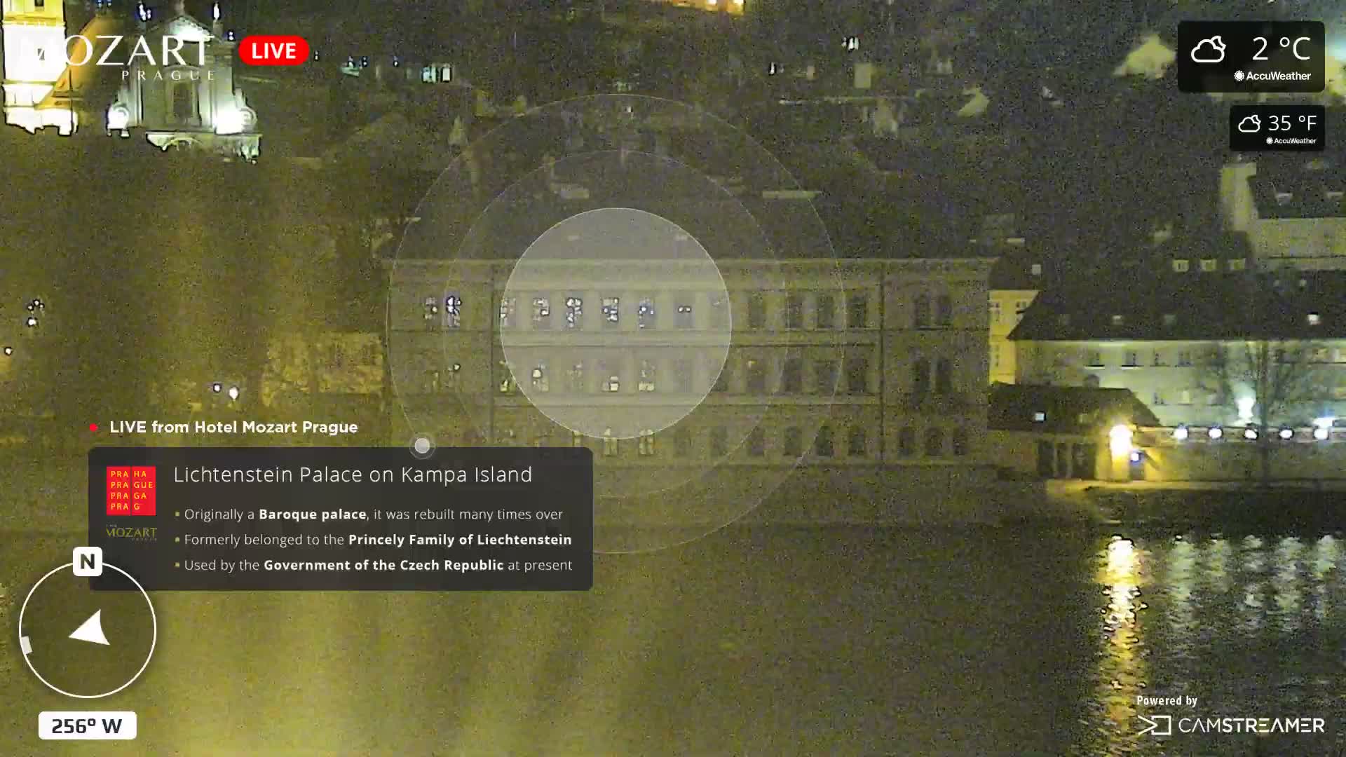 A grainy, nighttime view captures illuminated buildings and a church across a reflective body of water, with lights reflecting on the surface, under cloudy 2°C (35°F) conditions.